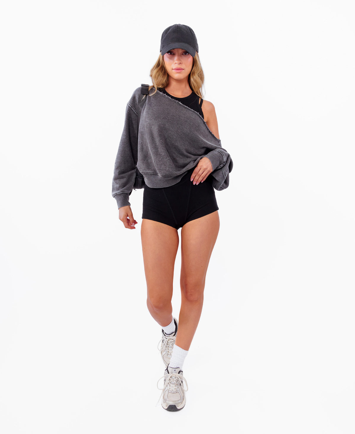 A model wearing a dark gray oversized off shoulder sweatshirt, black athletic shorts, a black cap, white socks, and gray sneakers, standing against a plain white background.