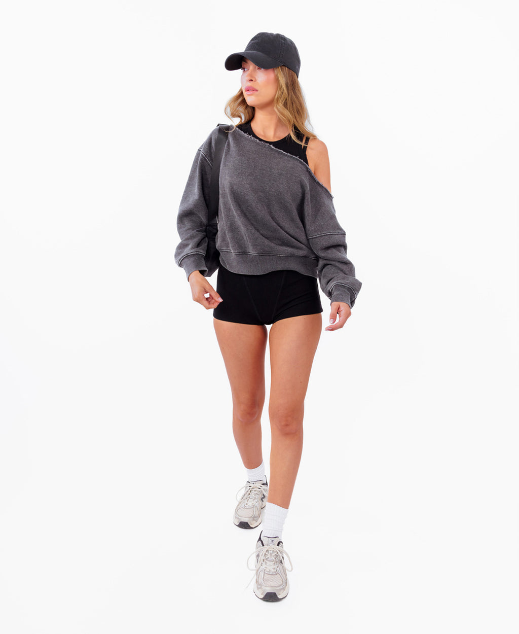 A model wearing a dark gray oversized off shoulder sweatshirt, black athletic shorts, a black cap, white socks, and gray sneakers, standing against a plain white background.
