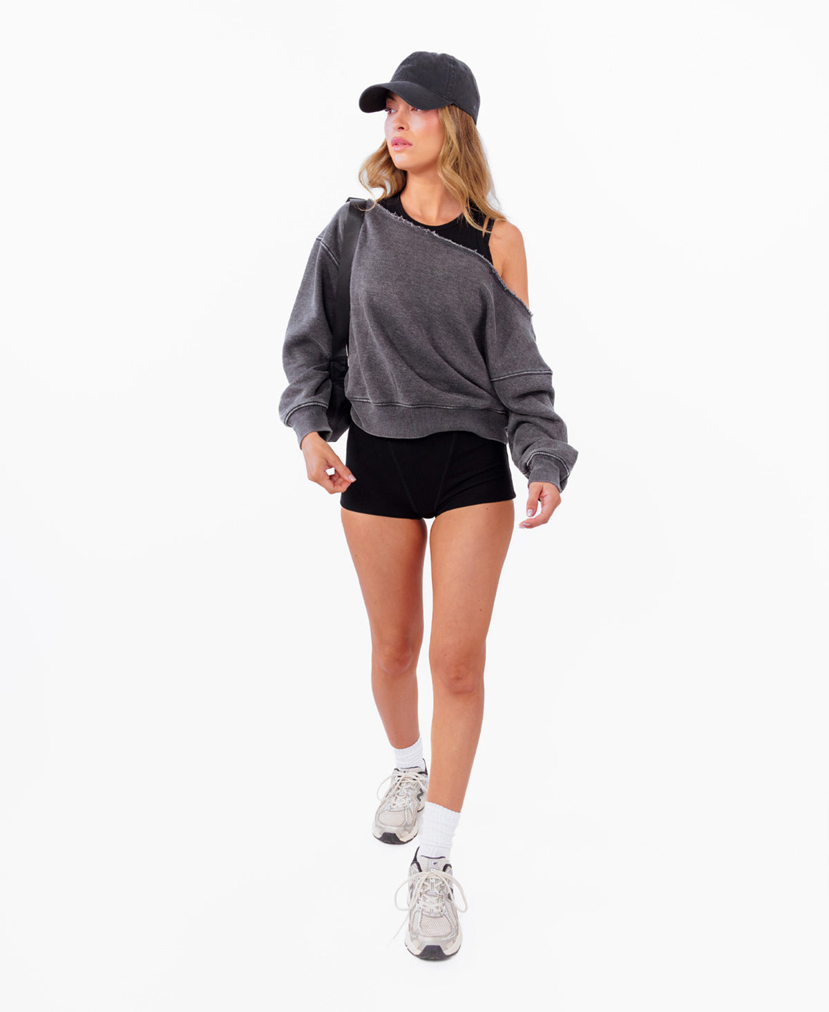 A model wearing a dark gray oversized off shoulder sweatshirt, black athletic shorts, a black cap, white socks, and gray sneakers, standing against a plain white background.