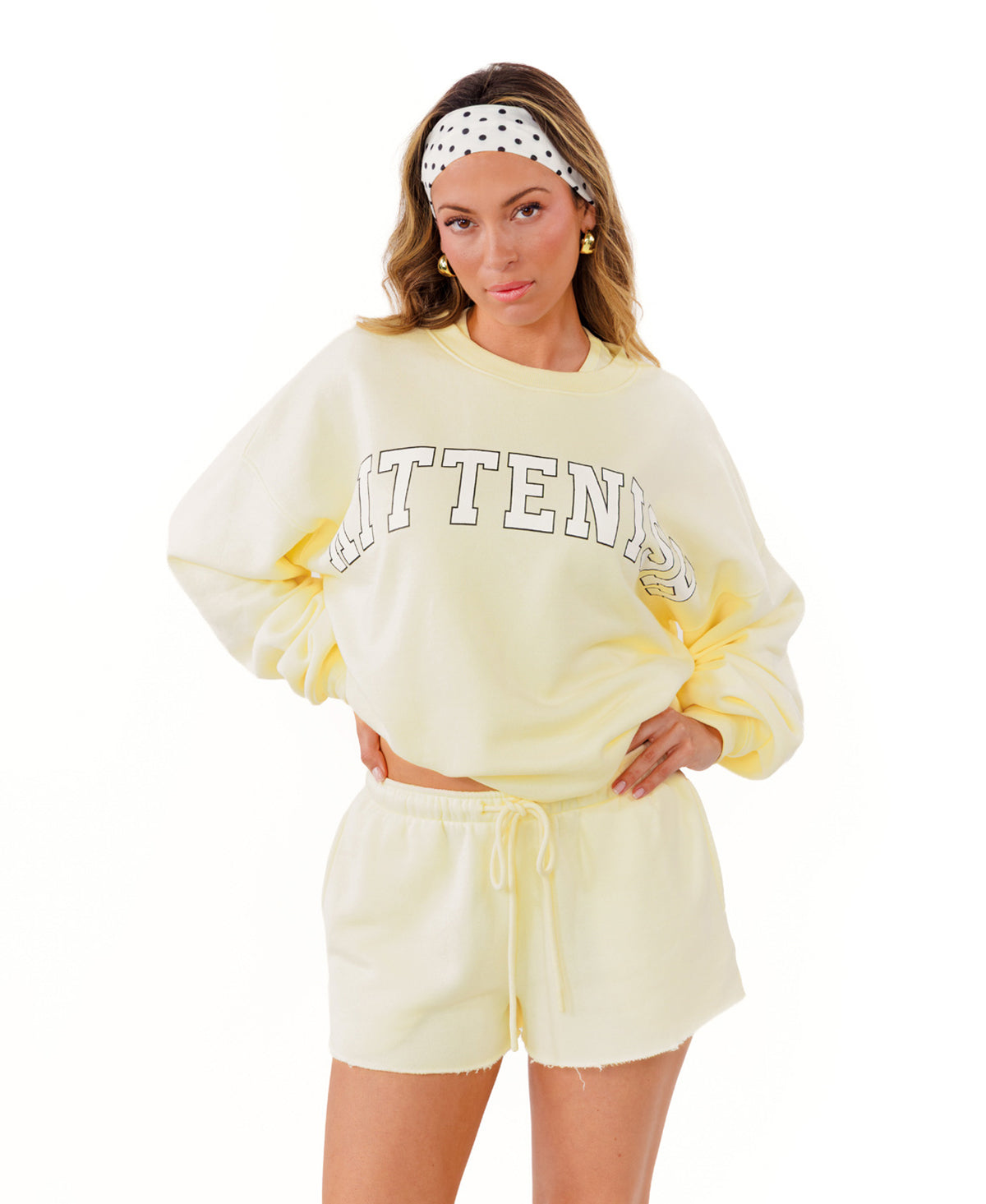 A woman modeling a pale yellow sweatshirt and matching shorts set against a plain white background. The sweatshirt features the word 'KITTENISH' in a white arched font outlined in black across the chest.
