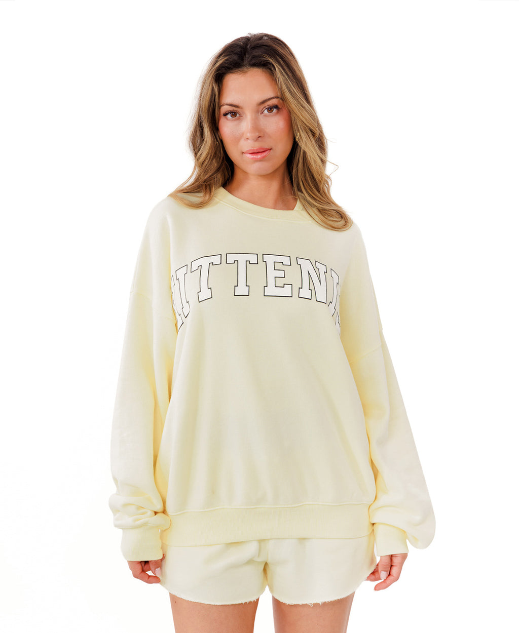 A woman modeling a butter yellow lounge set consisting of a sweatshirt with 'KITTENISH' printed in white across the chest and matching shorts, styled with white socks and white sneakers against a plain white background.
