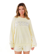 A woman modeling a butter yellow lounge set consisting of a sweatshirt with 'KITTENISH' printed in white across the chest and matching shorts, styled with white socks and white sneakers against a plain white background.