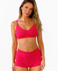 A woman modeling a bright pink bralette and boy shorts with horizontal bright pink and red stripes against a plain white background.