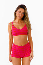 A woman modeling a bright pink bralette and boy shorts with horizontal bright pink and red stripes against a plain white background.