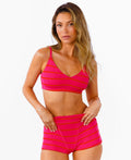 A woman modeling a bright pink bralette and boy shorts with horizontal bright pink and red stripes against a plain white background.