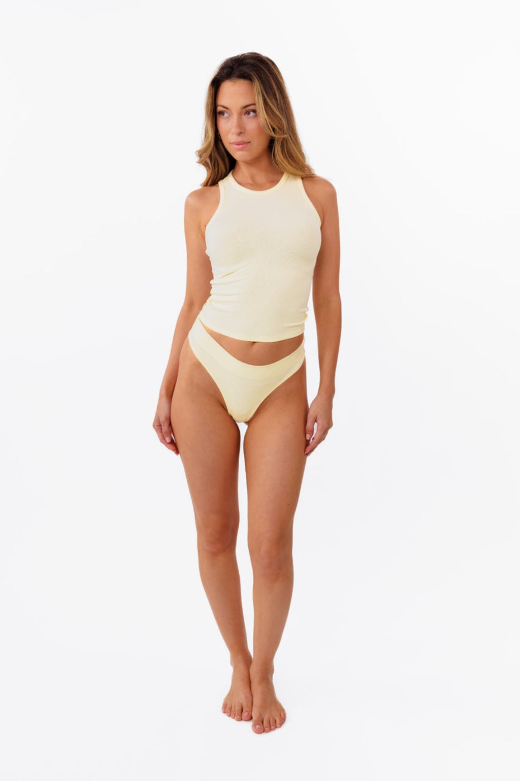 Model wearing pale yellow ribbed tank top and matching thong standing against a solid white background.