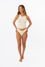 Model wearing pale yellow ribbed tank top and matching thong standing against a solid white background.
