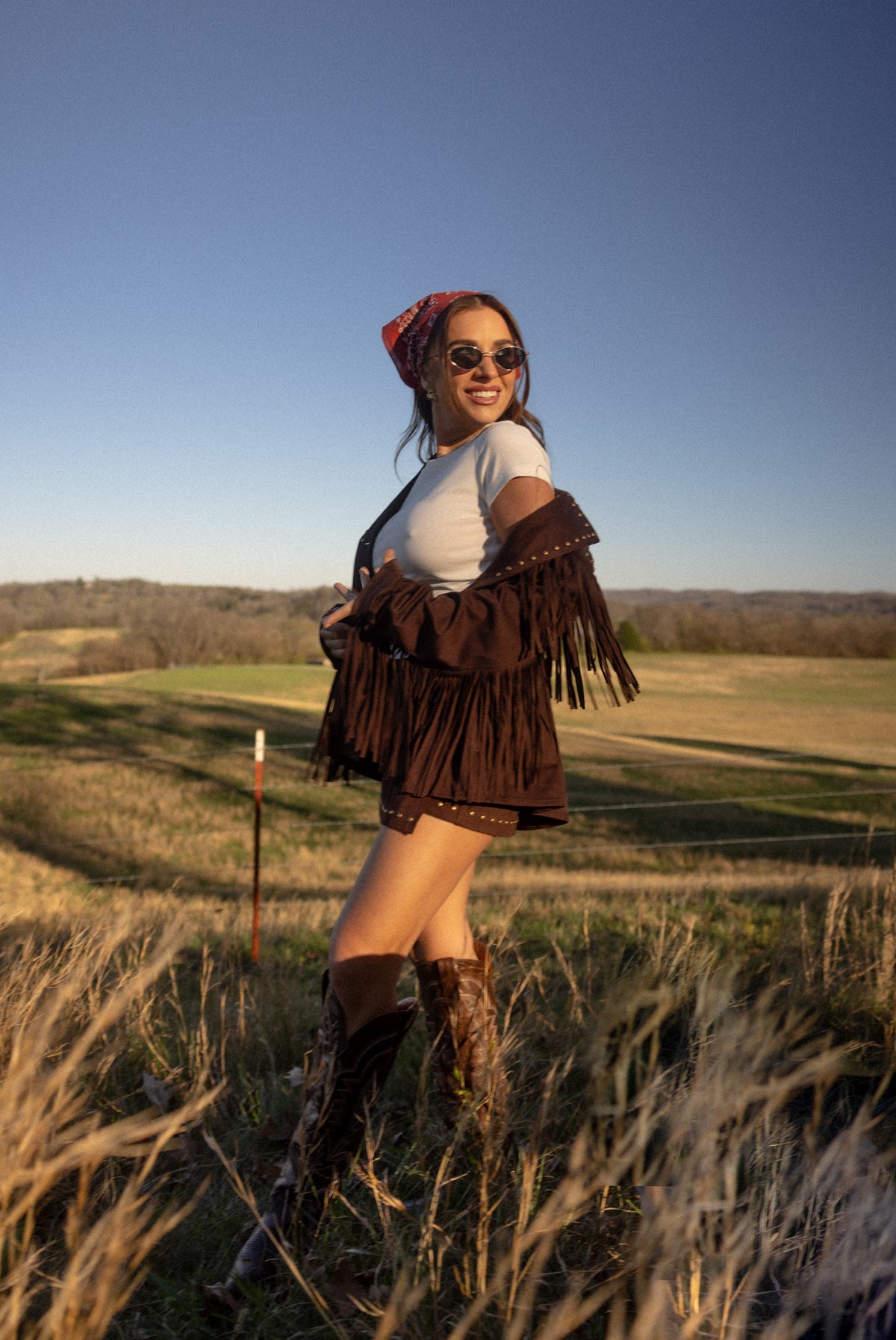Woman in a field wearing a brown fringe jacket, white top, and boots with a scenic background.