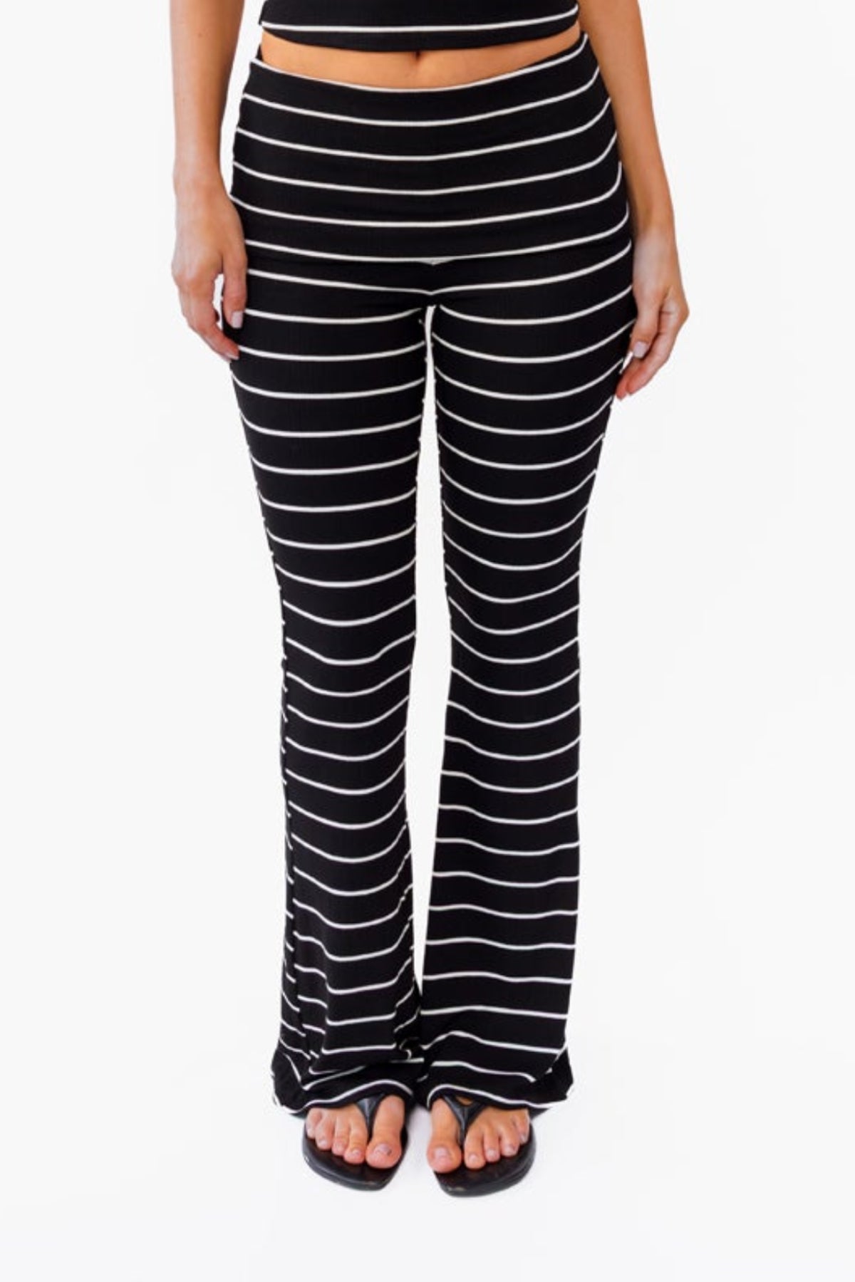 A woman modeling a black and white  two-piece lounge set with horizontal striped textures, consisting of a v neck tank top and matching fold over flared pant, against a plain white background.