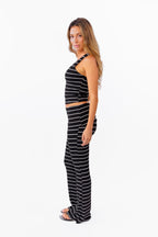 A woman modeling a black and white  two-piece lounge set with horizontal striped textures, consisting of a v neck tank top and matching fold over flared pant, against a plain white background.