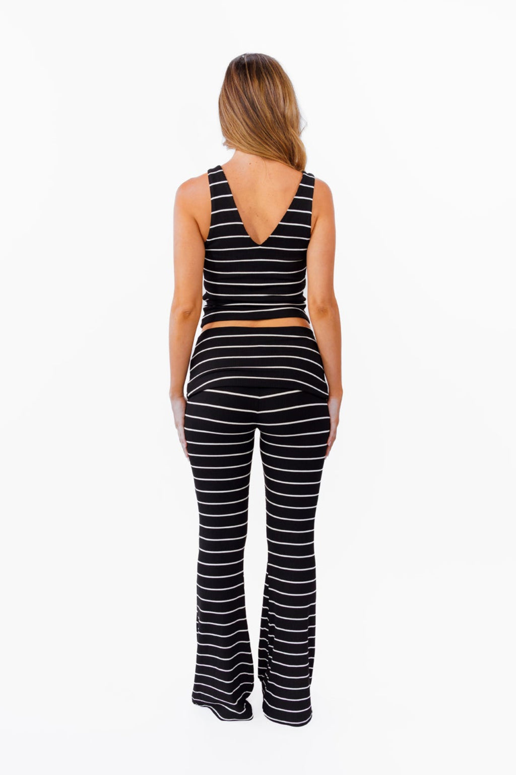 A woman modeling a black and white  two-piece lounge set with horizontal striped textures, consisting of a v neck tank top and matching fold over flared pant, against a plain white background.