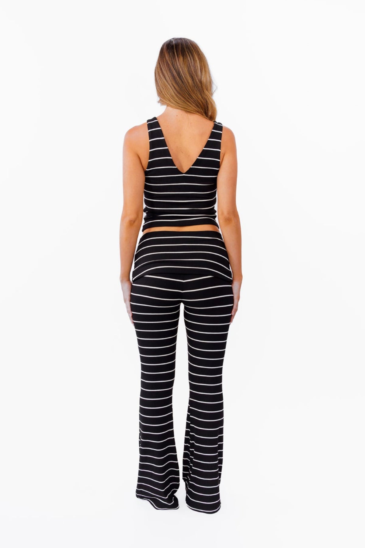 A woman modeling a black and white  two-piece lounge set with horizontal striped textures, consisting of a v neck tank top and matching fold over flared pant, against a plain white background.