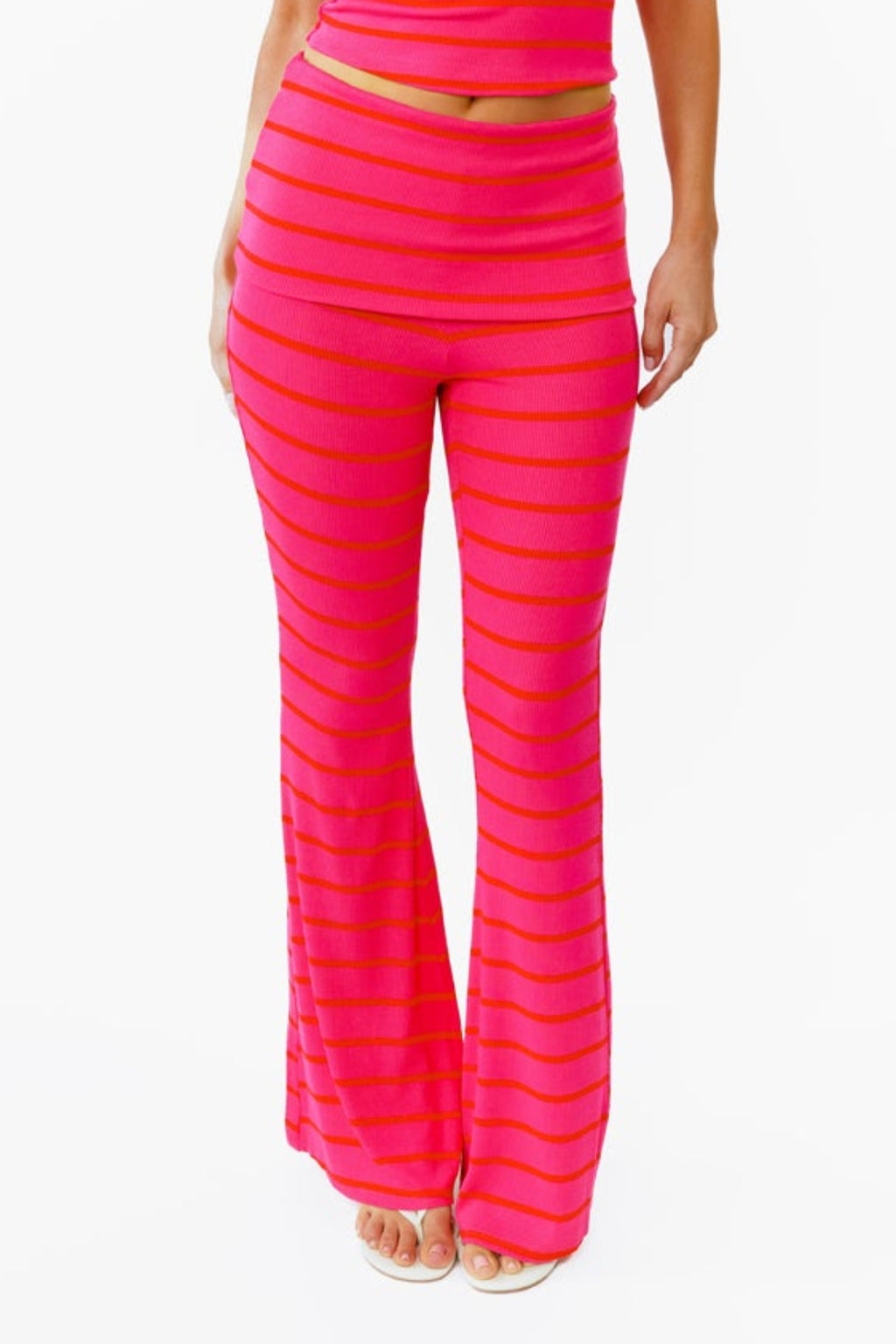 A woman modeling a bright pink two-piece lounge set with horizontal striped textures, consisting of a v neck tank top and matching flared pants with a fold over waistband, against a plain white background.