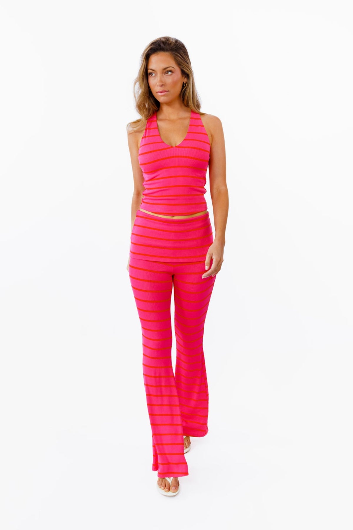 A woman modeling a bright pink two-piece lounge set with horizontal striped textures, consisting of a v neck tank top and matching flared pants with a fold over waistband, against a plain white background.
