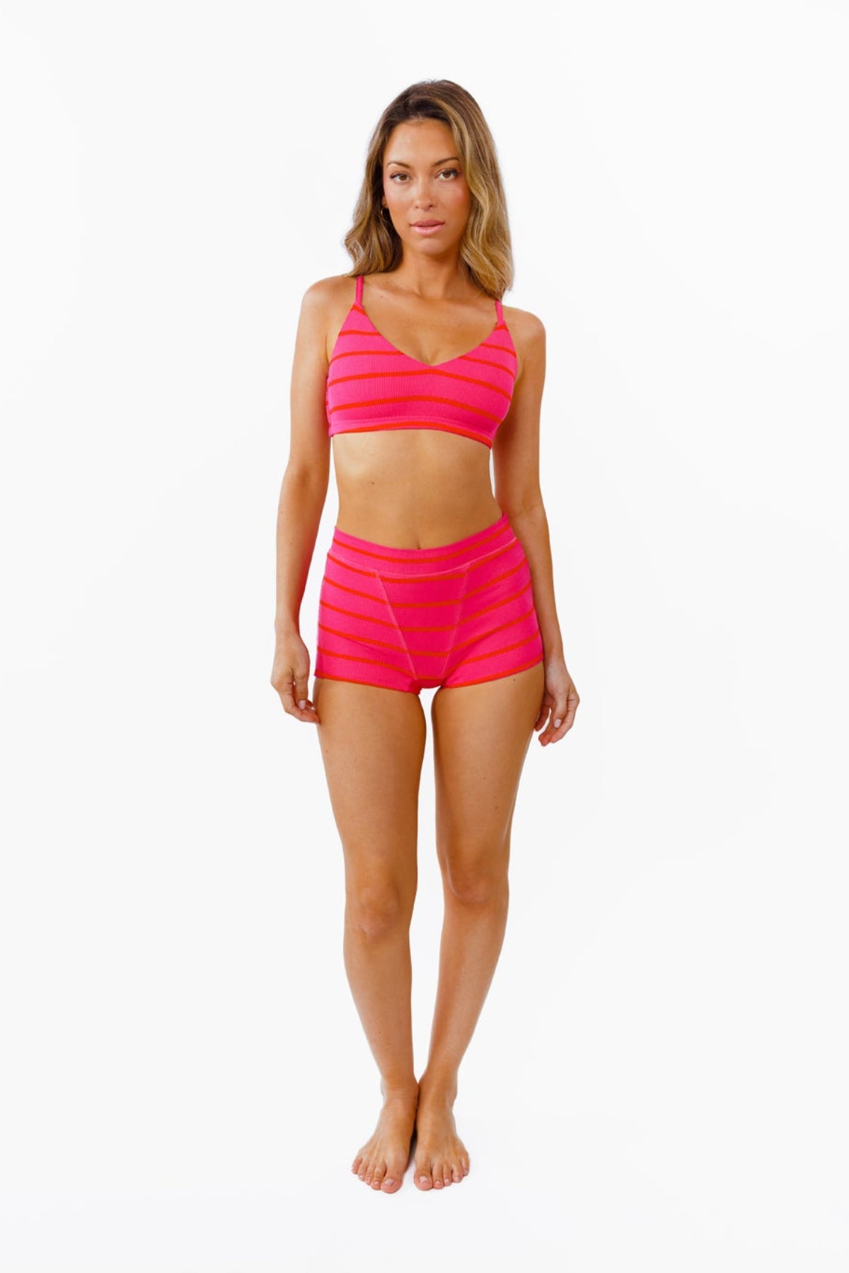 A woman modeling a bright pink bralette and boy shorts with horizontal bright pink and red stripes against a plain white background.