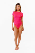 A woman modeling a bright pink and red  horizontal stripe thong and short sleeve top standing against a plain white background.
