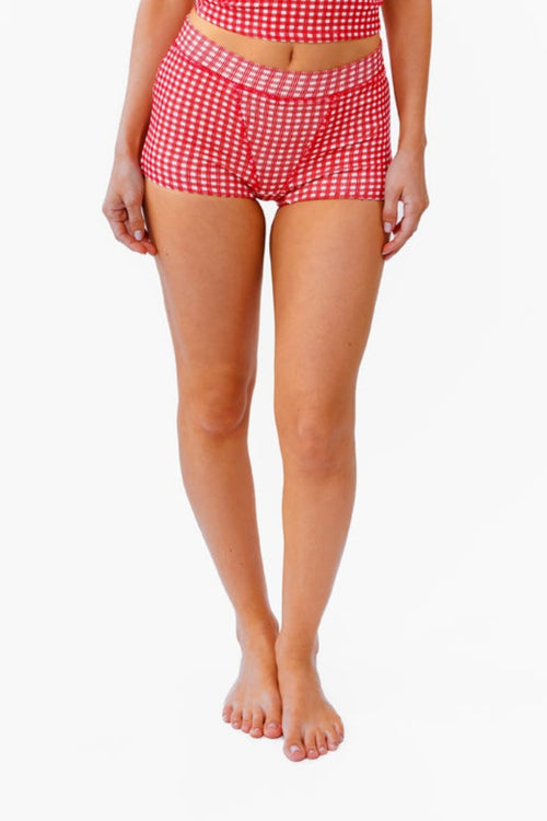 A woman modeling a two-piece red and white gingham set consisting of a sleeveless crop tank top and matching high-waisted boy shorts, standing against a plain white background.
