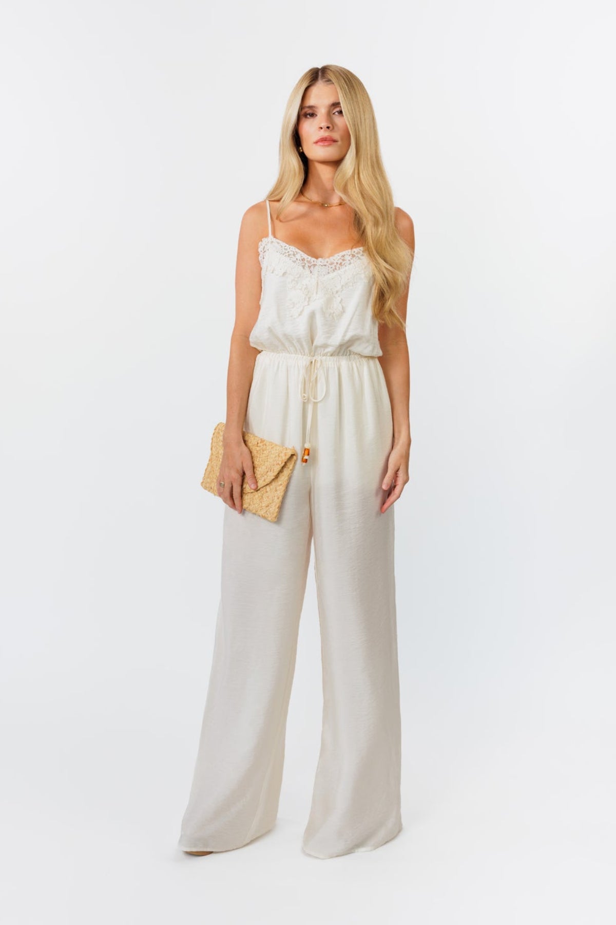 Woman wearing a white lace wide-leg jumpsuit holding a straw clutch against a white background