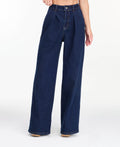 A model wearing dark blue wide-leg denim jeans with a high waist and front pleats, shown from the waist down on a white studio background.