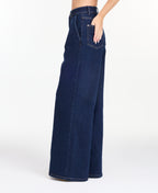A model wearing dark blue wide-leg denim jeans with a high waist and front pleats, shown from the waist down on a white studio background.