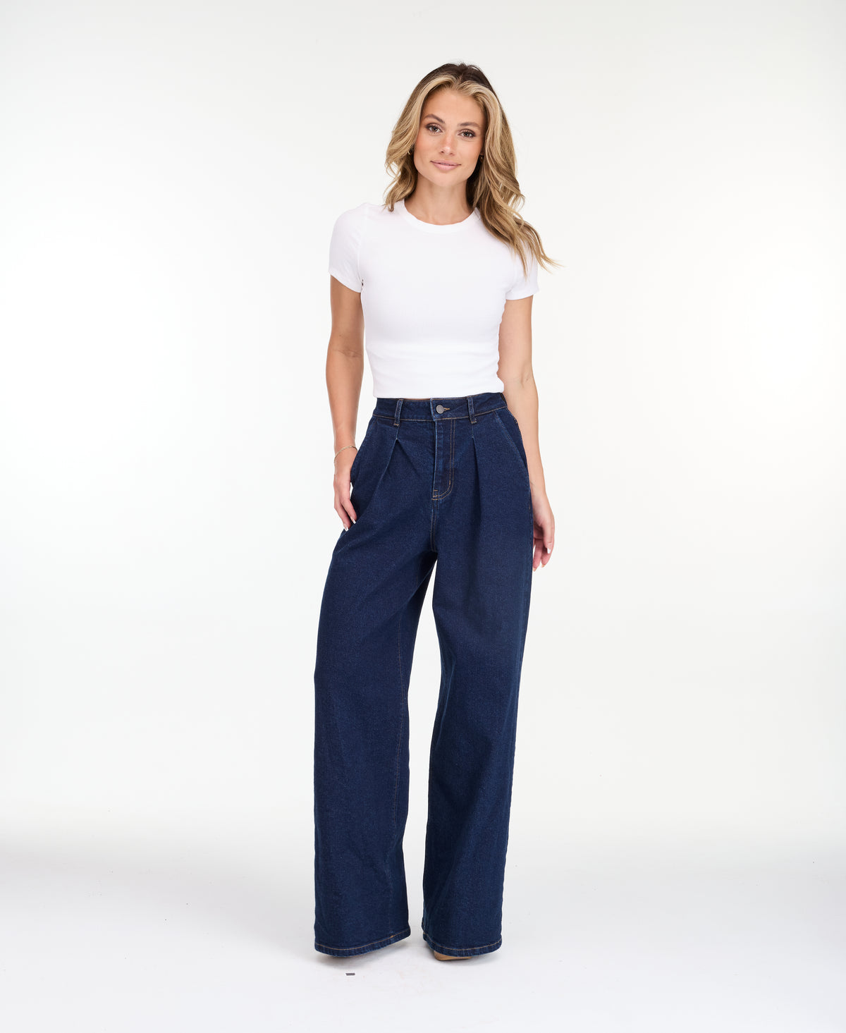 A model wearing dark blue wide-leg denim jeans with a high waist and front pleats, shown from the waist down on a white studio background.