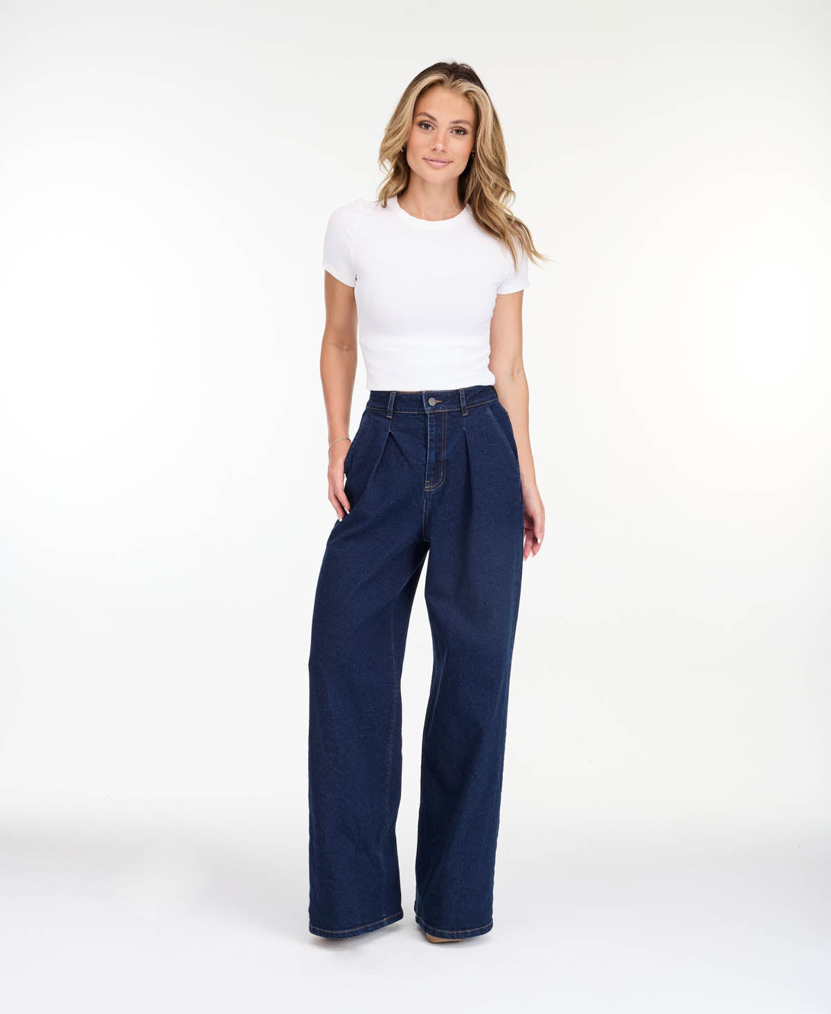 A model wearing dark blue wide-leg denim jeans with a high waist and front pleats, shown from the waist down on a white studio background.