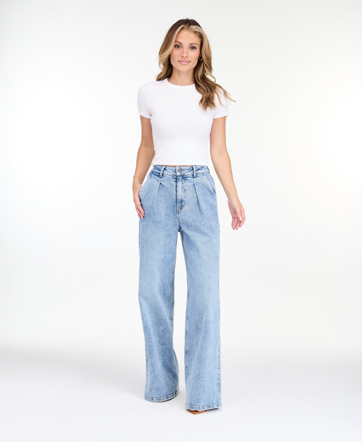 A woman modeling a light-wash blue denim outfit consisting of wide-leg jeans, a white ribbed top, and an open light blue denim jacket, photographed against a seamless white studio background.