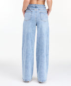 A woman modeling a light-wash blue denim outfit consisting of wide-leg jeans, a white ribbed top, and an open light blue denim jacket, photographed against a seamless white studio background.