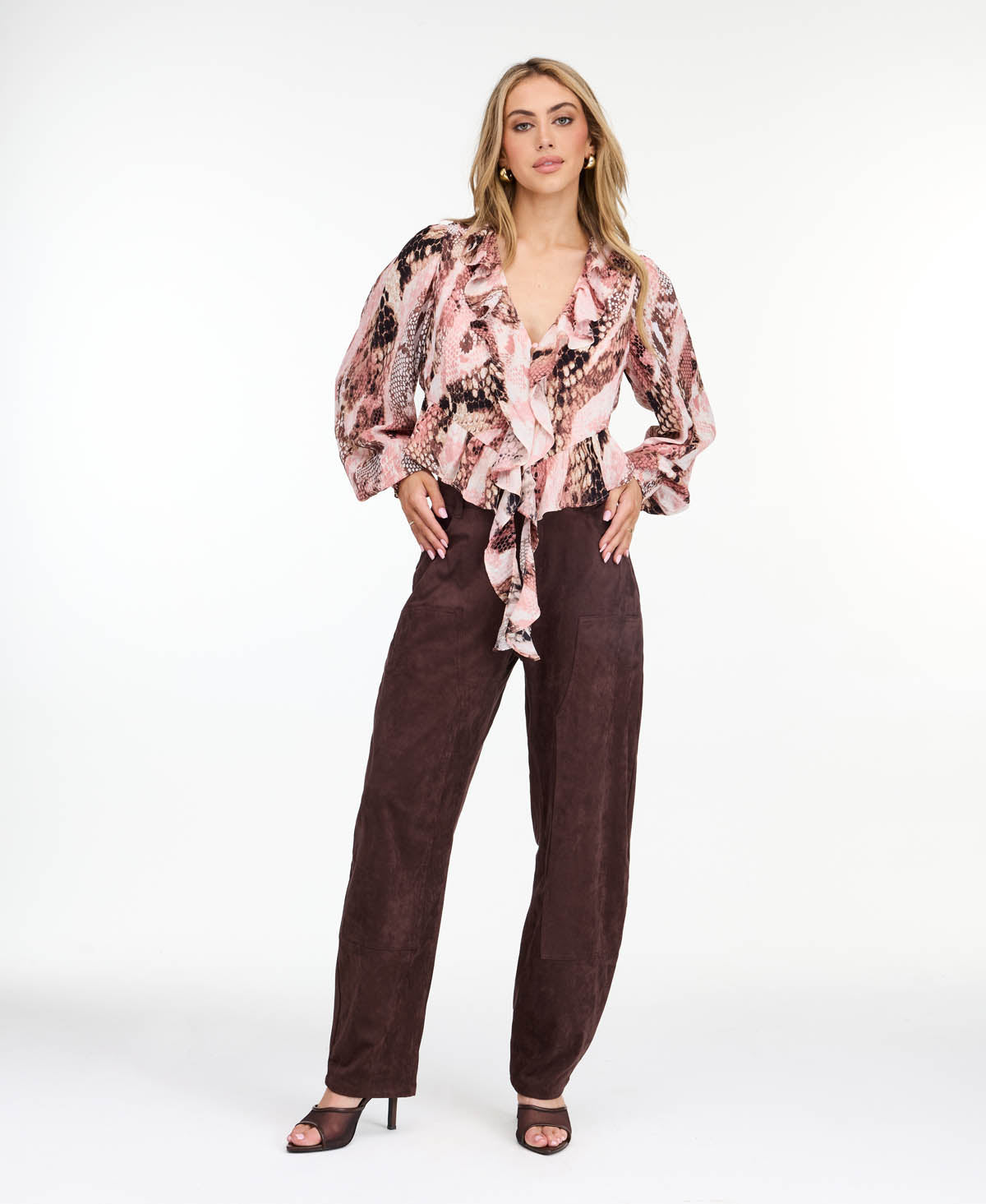 Model wearing brown faux suede barrel pants with brown kitten heels and a snake print top standing in front of a white back drop