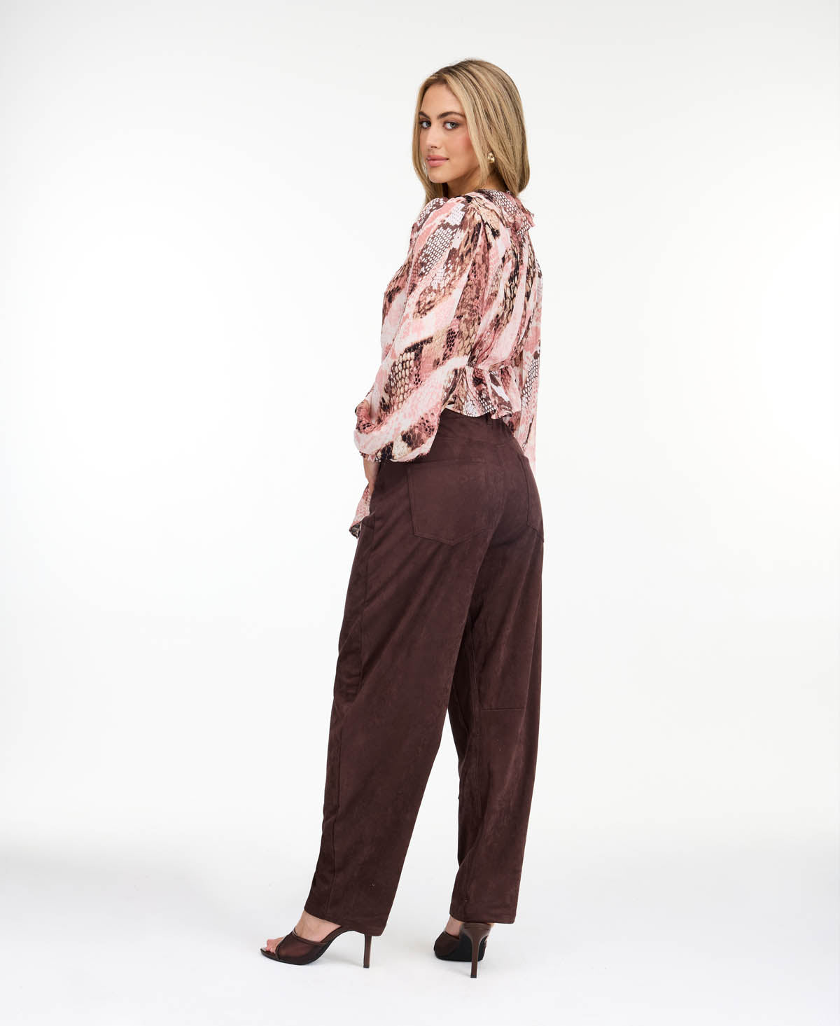 Model wearing brown faux suede barrel pants with brown kitten heels and a snake print top standing in front of a white back drop
