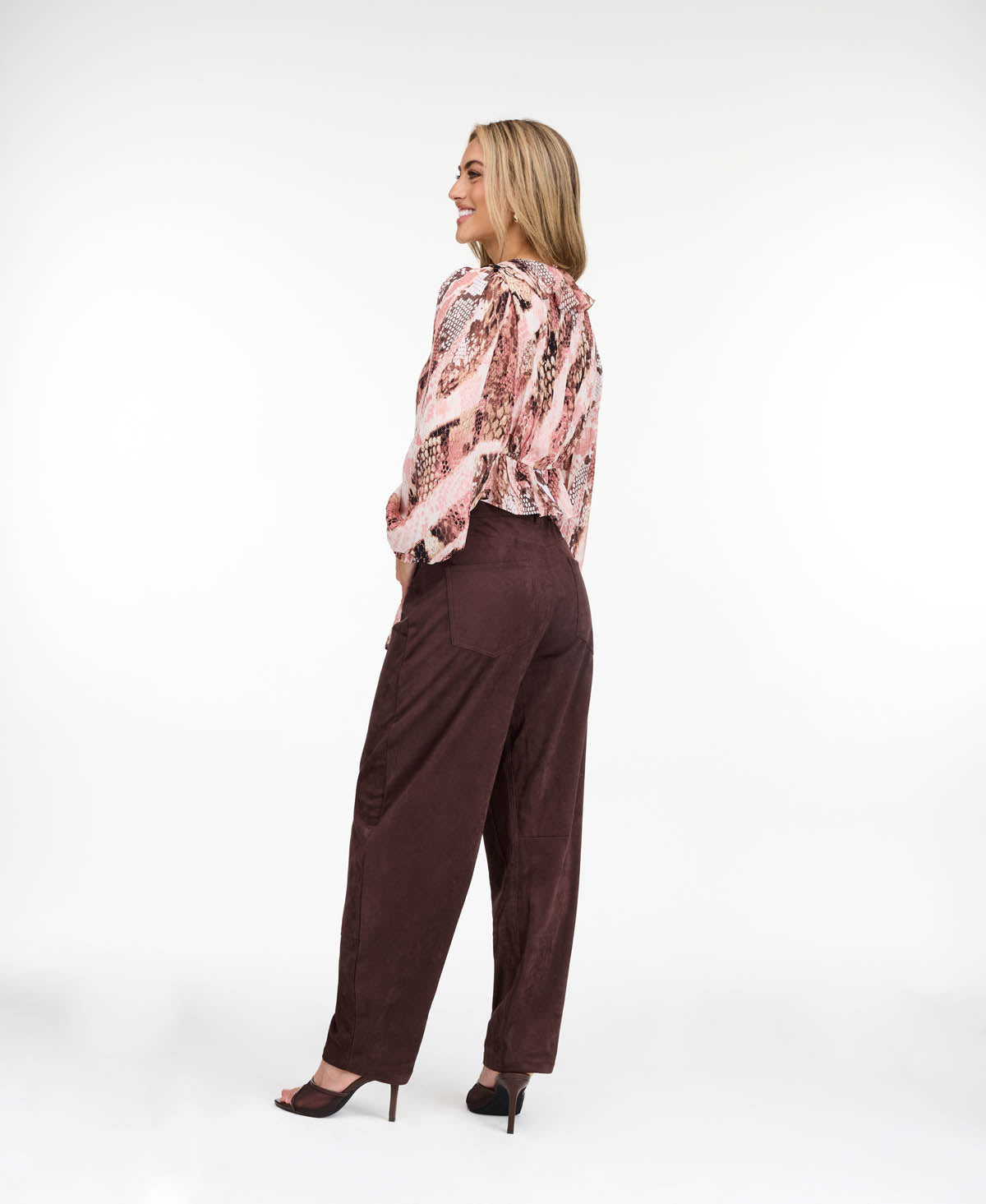 Model wearing a pink snake print top with brown suede pants and brown kitten heels standing against a white back drop