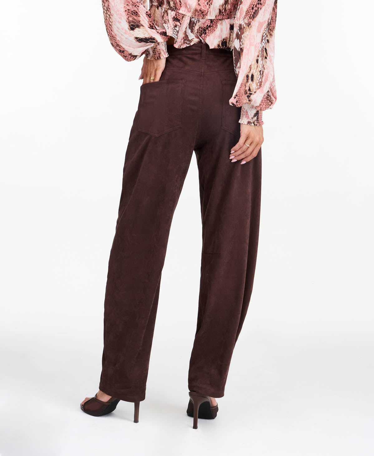 Model wearing brown faux suede barrel pants with brown kitten heels and a snake print top standing in front of a white back drop