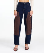 A woman modeling a dark brown fringed jacket with studded detailing, paired with dark brown and navy patchwork pants and brown heels, against a plain white studio background.