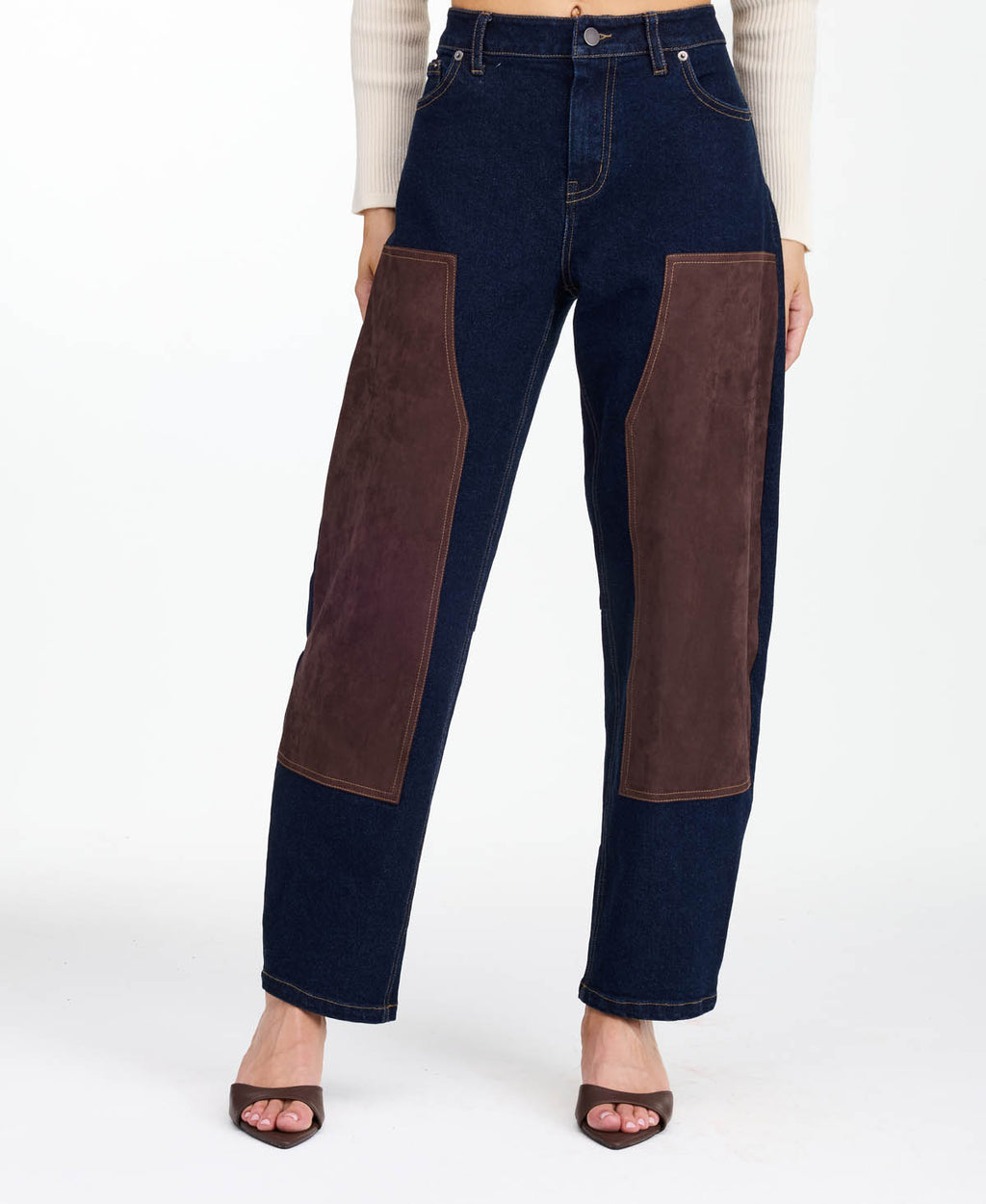 A woman modeling a dark brown fringed jacket with studded detailing, paired with dark brown and navy patchwork pants and brown heels, against a plain white studio background.