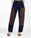 A woman modeling a dark brown fringed jacket with studded detailing, paired with dark brown and navy patchwork pants and brown heels, against a plain white studio background.