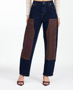 A woman modeling a dark brown fringed jacket with studded detailing, paired with dark brown and navy patchwork pants and brown heels, against a plain white studio background.