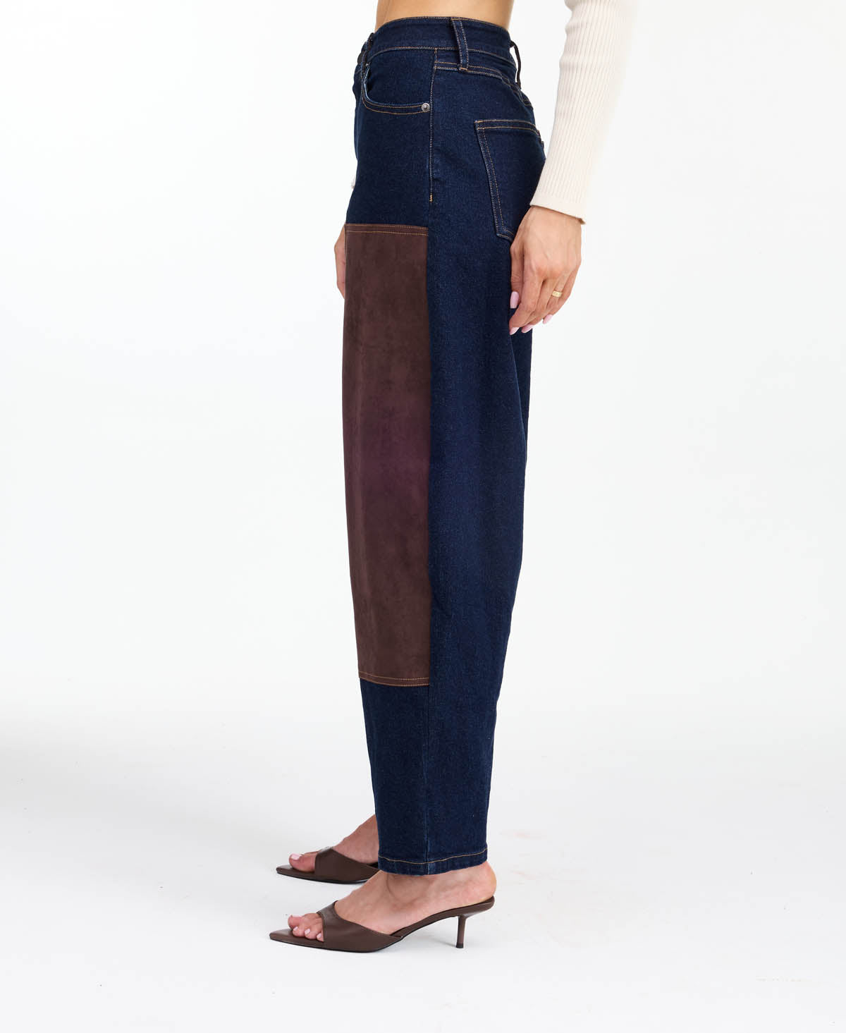 A woman modeling a dark brown fringed jacket with studded detailing, paired with dark brown and navy patchwork pants and brown heels, against a plain white studio background.