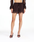 A model wearing dark brown lace shorts with a drawstring waist and lace trim edges, paired with matching brown heeled sandals and a matching top, against a plain white studio background.