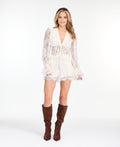 A woman modeling a white lace long-sleeve top with a deep V-neck and ruffled hem and matching shorts, paired with brown suede knee-high boots, against a plain white studio background.