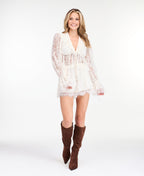 A woman modeling a white lace long-sleeve top with a deep V-neck and ruffled hem and matching shorts, paired with brown suede knee-high boots, against a plain white studio background.