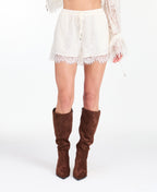 A woman modeling a white lace long-sleeve mini dress with a deep V-neck and ruffled hem, paired with brown suede knee-high boots, against a plain white studio background.