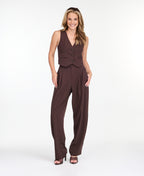 A model is shown from the waist down wearing high-waisted, wide-leg brown trousers with thin vertical pinstripes, paired with brown  heels against a plain white studio background.