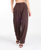 A model is shown from the waist down wearing high-waisted, wide-leg brown trousers with thin vertical pinstripes, paired with brown  heels against a plain white studio background.