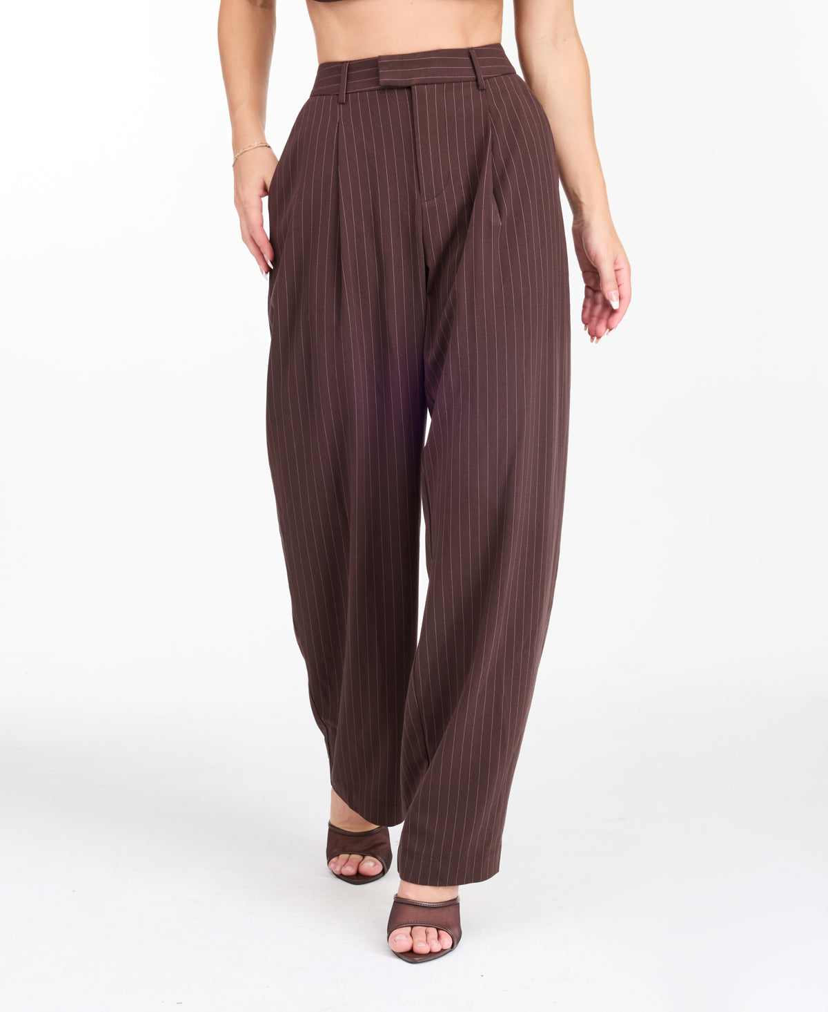 A model is shown from the waist down wearing high-waisted, wide-leg brown trousers with thin vertical pinstripes, paired with brown  heels against a plain white studio background.