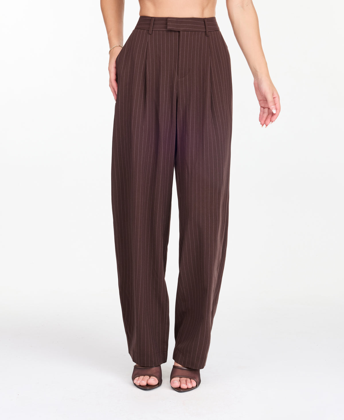 A model is shown from the waist down wearing high-waisted, wide-leg brown trousers with thin vertical pinstripes, paired with brown  heels against a plain white studio background.