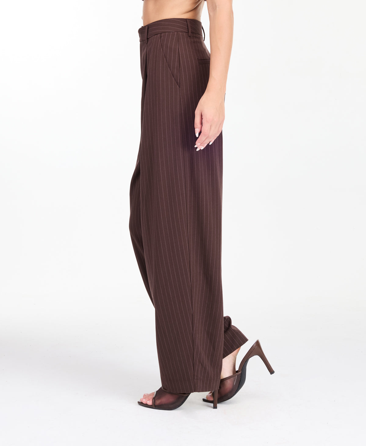 A model is shown from the waist down wearing high-waisted, wide-leg brown trousers with thin vertical pinstripes, paired with brown  heels against a plain white studio background.