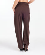 A model is shown from the waist down wearing high-waisted, wide-leg brown trousers with thin vertical pinstripes, paired with brown  heels against a plain white studio background.