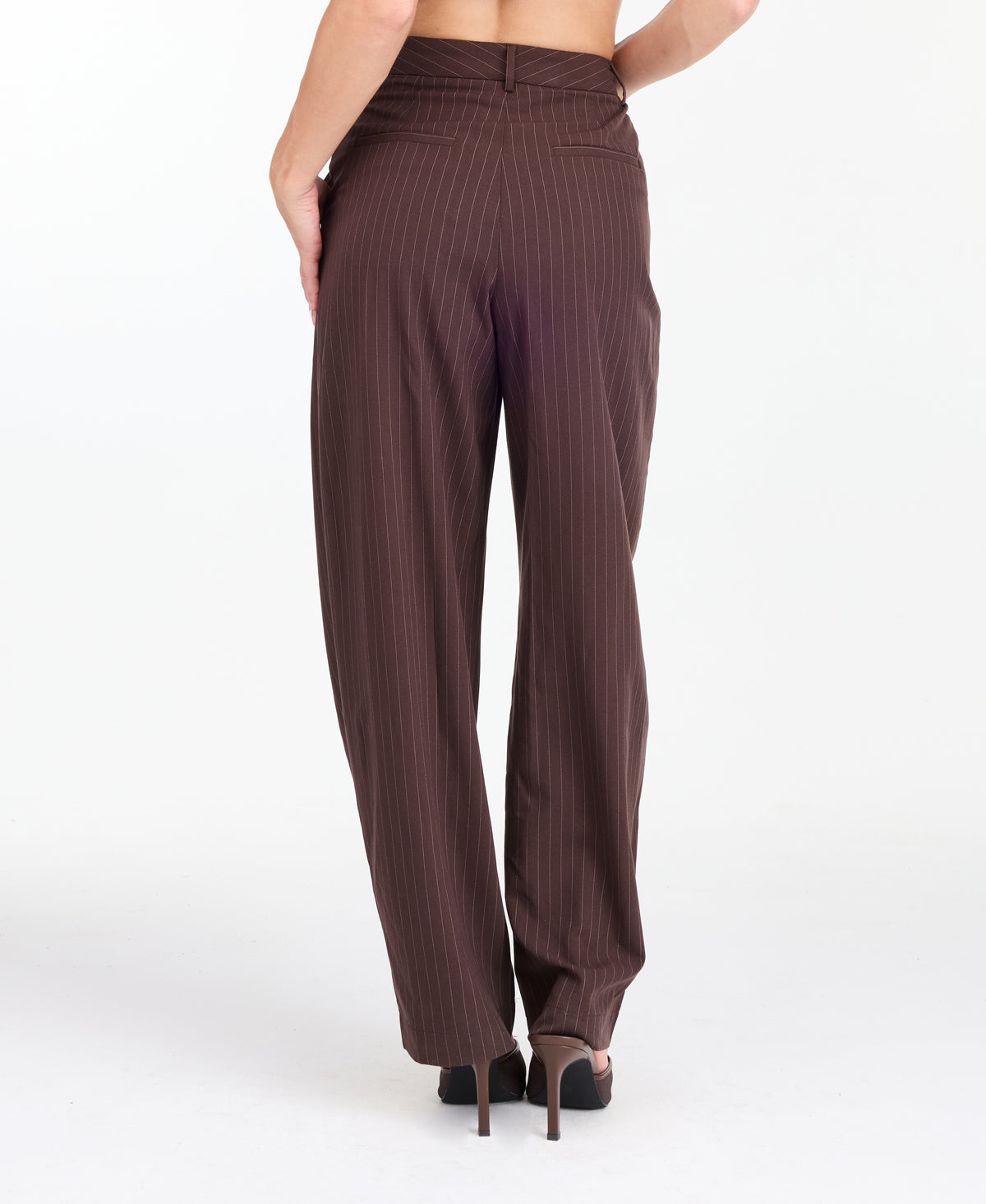 A model is shown from the waist down wearing high-waisted, wide-leg brown trousers with thin vertical pinstripes, paired with brown  heels against a plain white studio background.