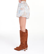 A model wearing a high-waisted bubble shorts and a matching top with a light blue, beige, and white abstract print, paired with brown suede knee-high boots, against a plain white studio background.