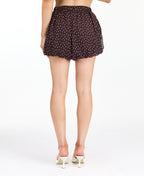 A model from the waist down wearing dark brown shorts with a white polka dot pattern, paired with white heeled sandals, against a plain white studio background.